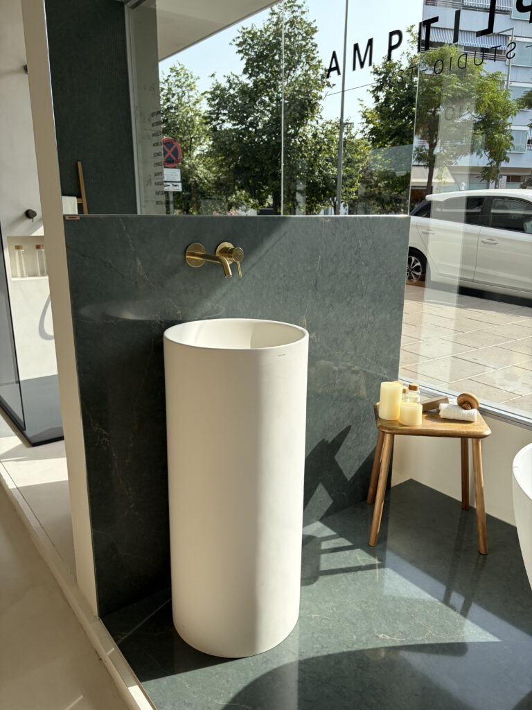 Lavabo exento cilíndrico con grifería dorada y mármol verde en el showroom de Amplitud Studio